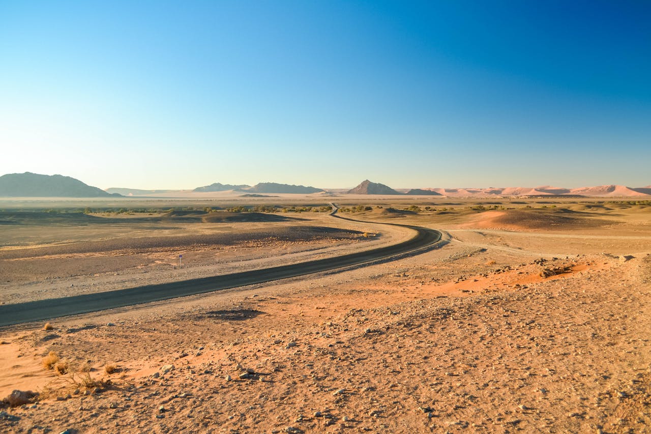 Home A winding road through the vast Namib Desert landscape under a clear blue sky.
